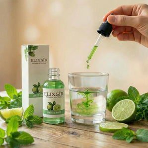 Elixsír Limette & Minze-Sirup: grüner Pipettentropfen im Wasserglas, daneben Flasche, Schachtel, Limetten und Minze