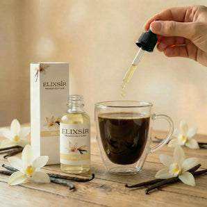 Elixsír Vanille-Sirup wird mit der Pipette in eine Glastasse Kaffee geträufelt, daneben Flasche, Vanilleblüten und -schoten